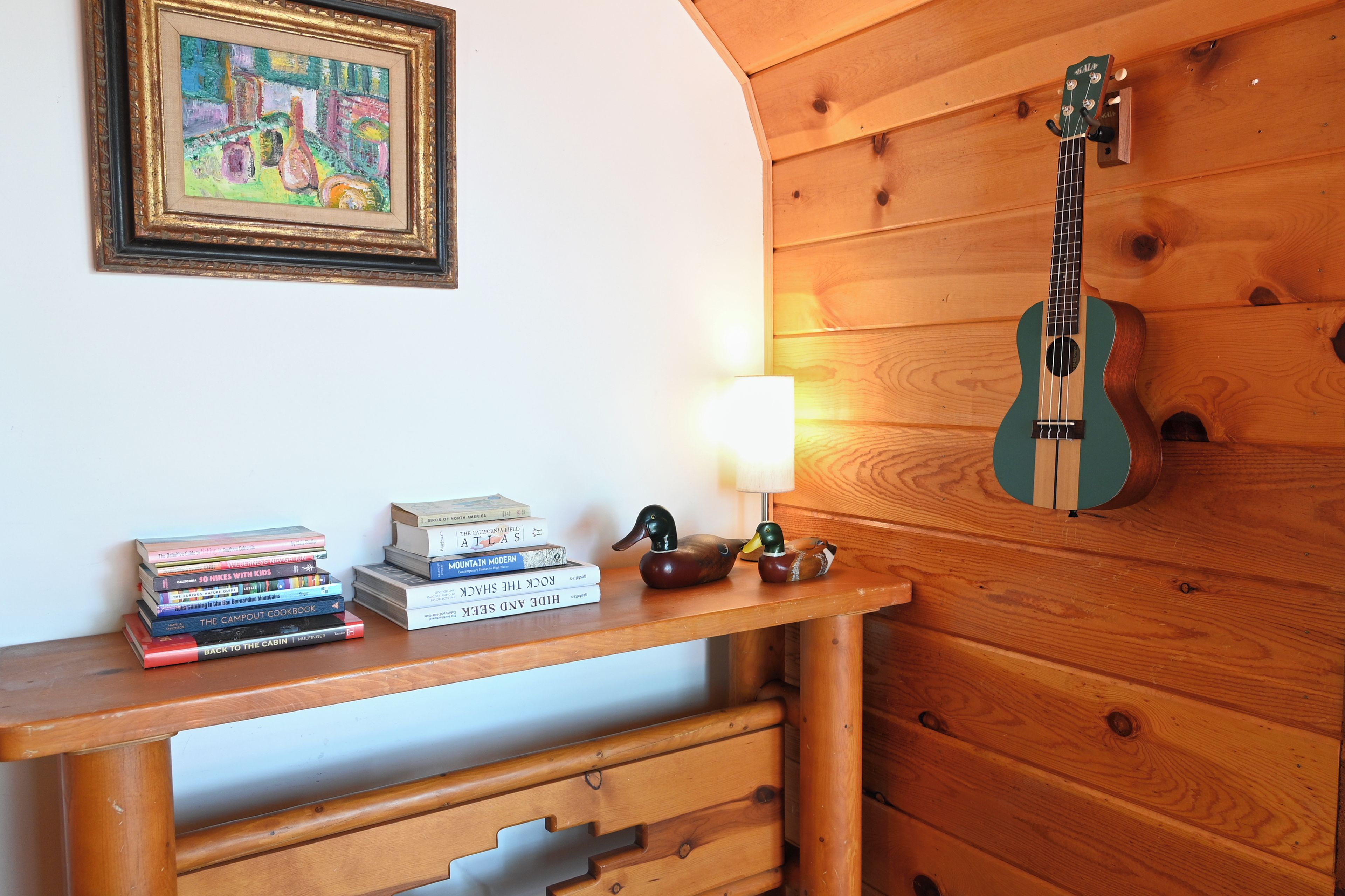 Eine Holzwand mit einer an ihr lehnten Gitarre, einem Bücherstapel und einer Lampe auf einem Regal.