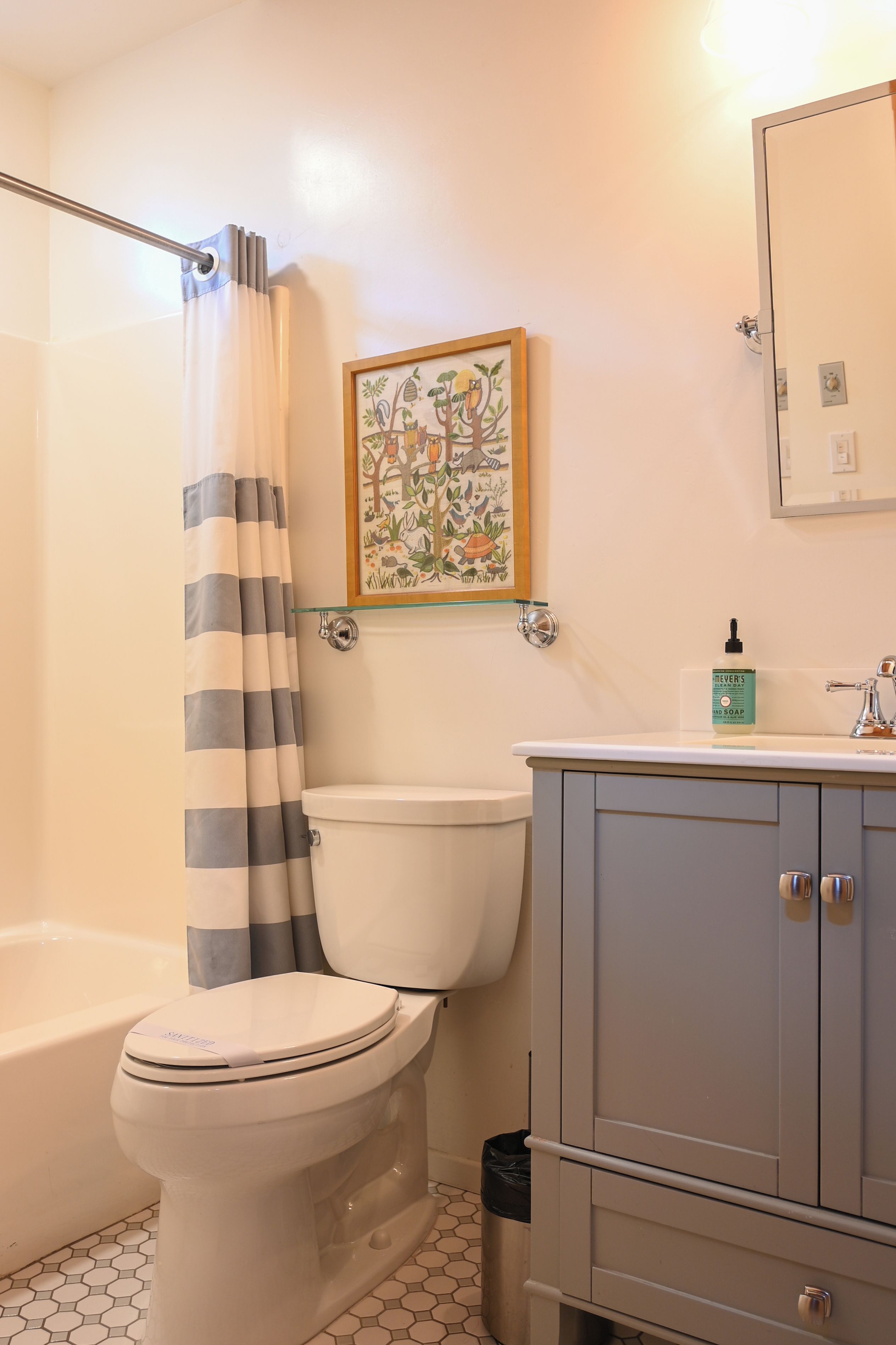 Une salle de bain comprenant des toilettes, un rideau de douche, un tableau encadré au mur, un lavabo avec un distributeur de savon et un meuble de rangement.