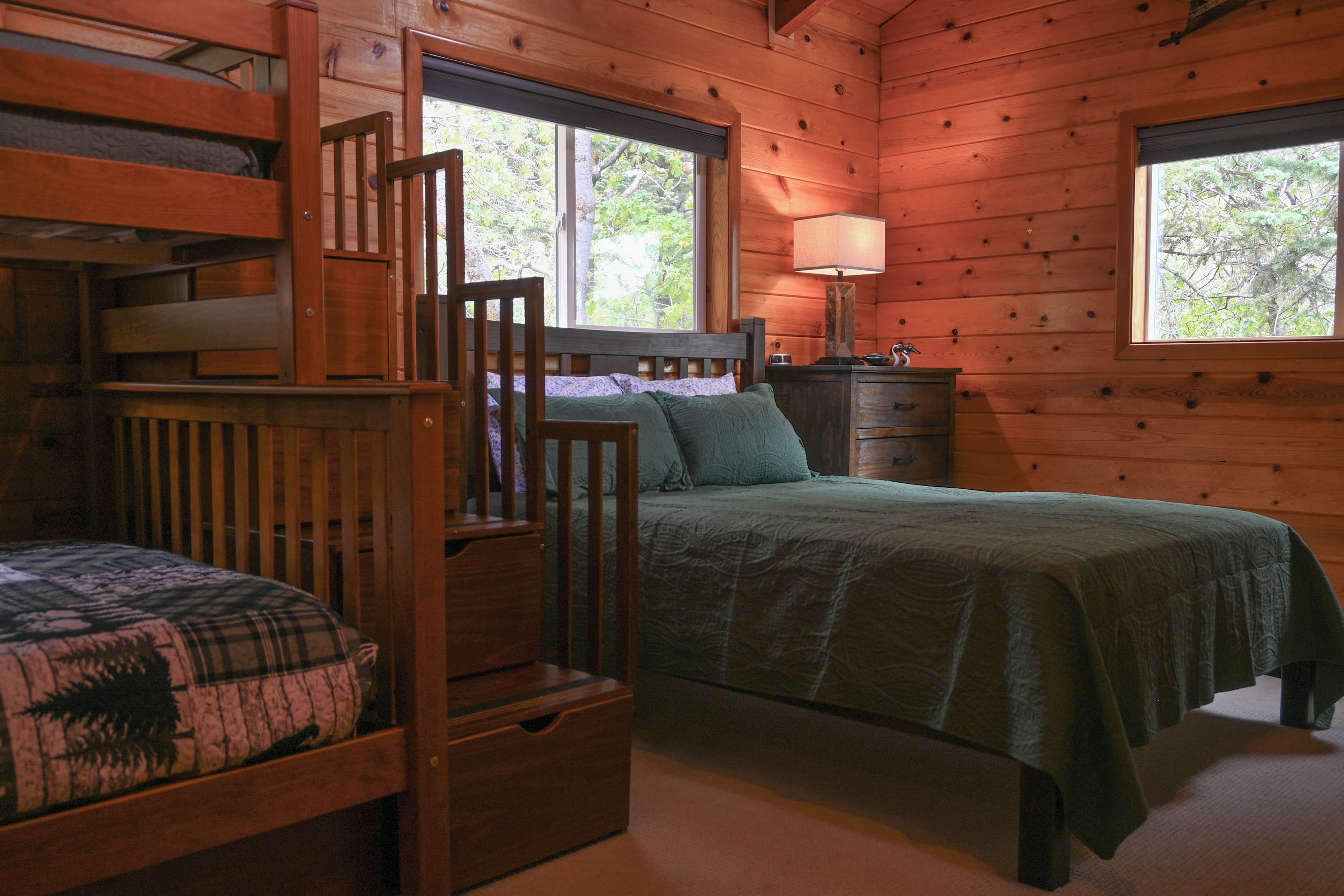 A cabin interior with a bunk bed, a single bed, a dresser, and a window with a view of trees.