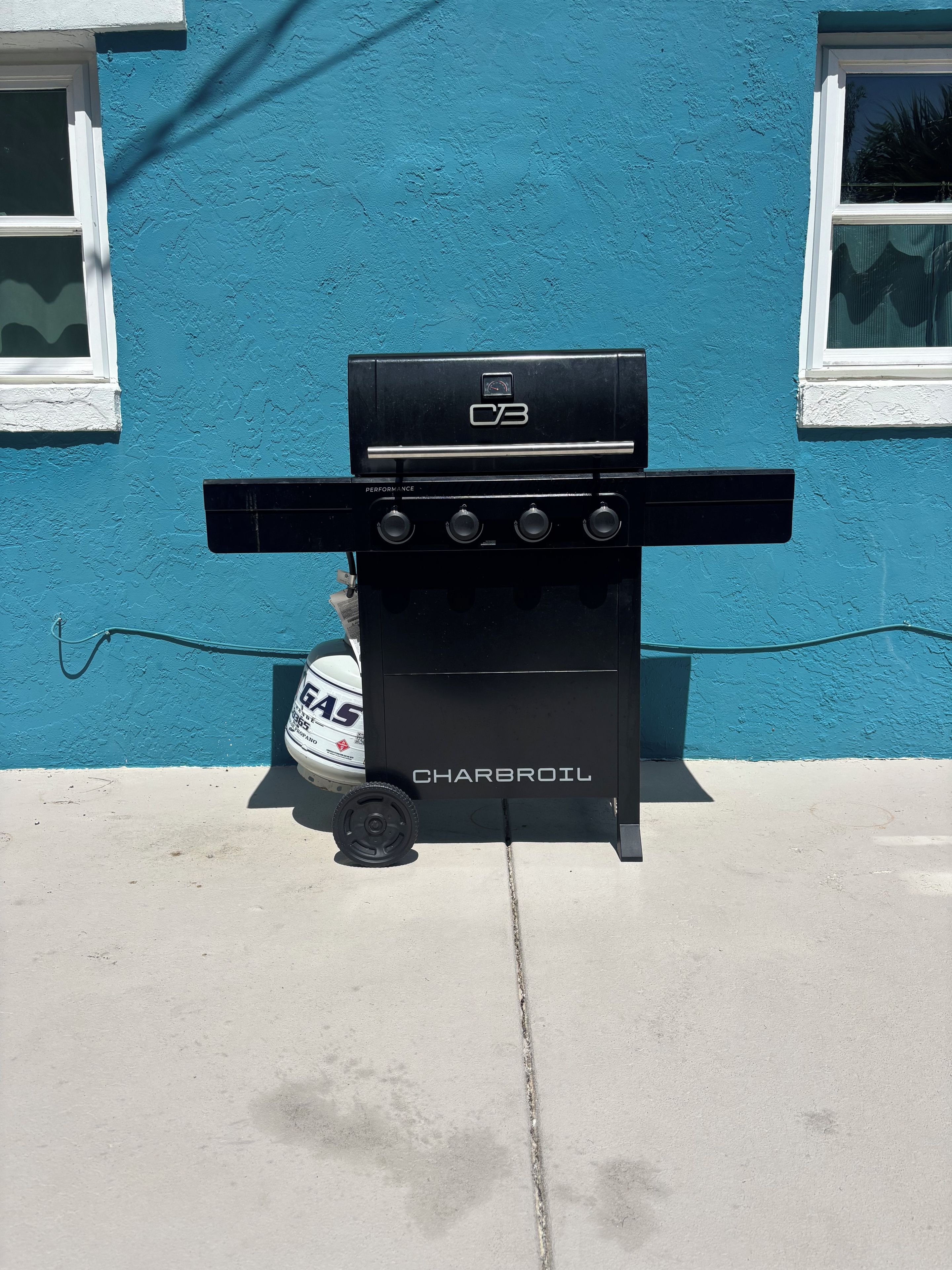Una parrilla Charbroil negra con cuatro quemadores, ubicada frente a una casa azul con marcos de ventana blancos.