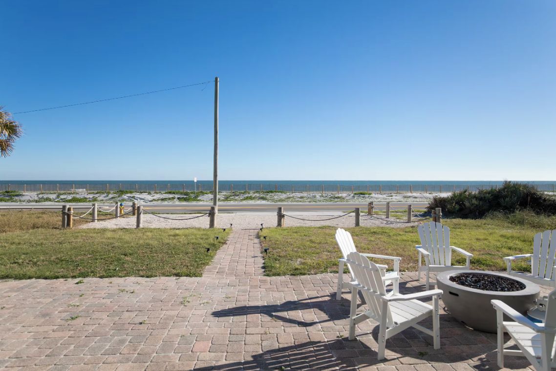 A paved pathway leading to beach chairs and a fire pit, overlooking a sandy beach with a clear sky.