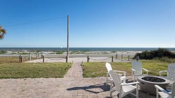 A paved pathway leading to beach chairs and a fire pit, overlooking a sandy beach with a clear sky.