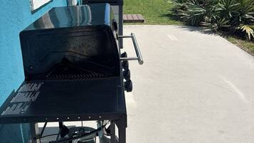 A barbecue grill with a propane tank, a blue building, a palm tree, and a clear sky.
