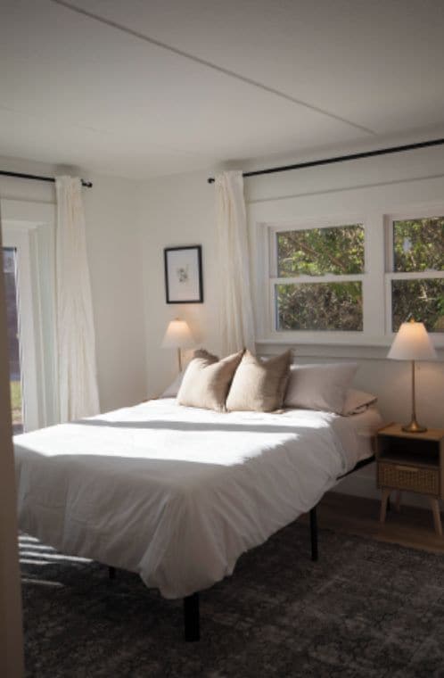 A bedroom with a bed, two bedside lamps, a window with curtains, and a framed picture on the wall.