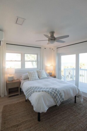Un dormitorio con cama, mesitas de noche, ventilador de techo y ventanas grandes.