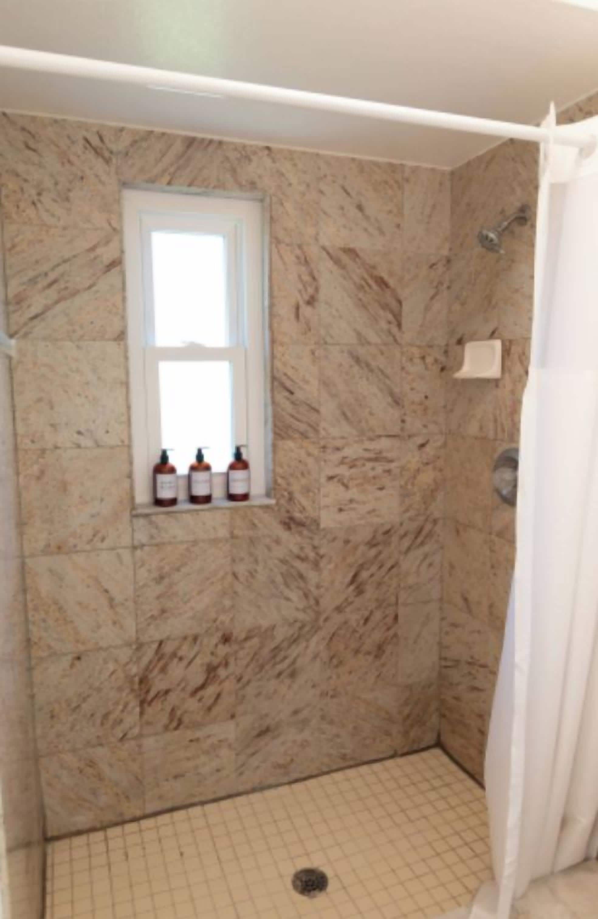 A bathroom with a shower, a window, and a shelf with toiletries.