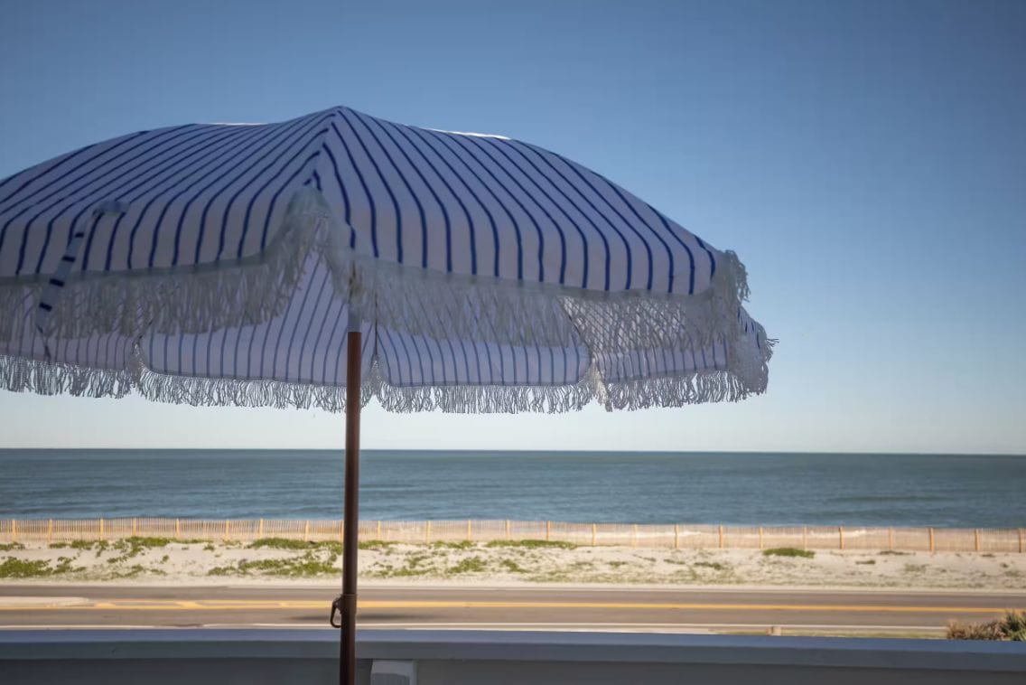 Un parasol de plage à motifs rayés et à franges, placé au-dessus d’une route d’où l’on peut admirer l’océan et la plage de sable.