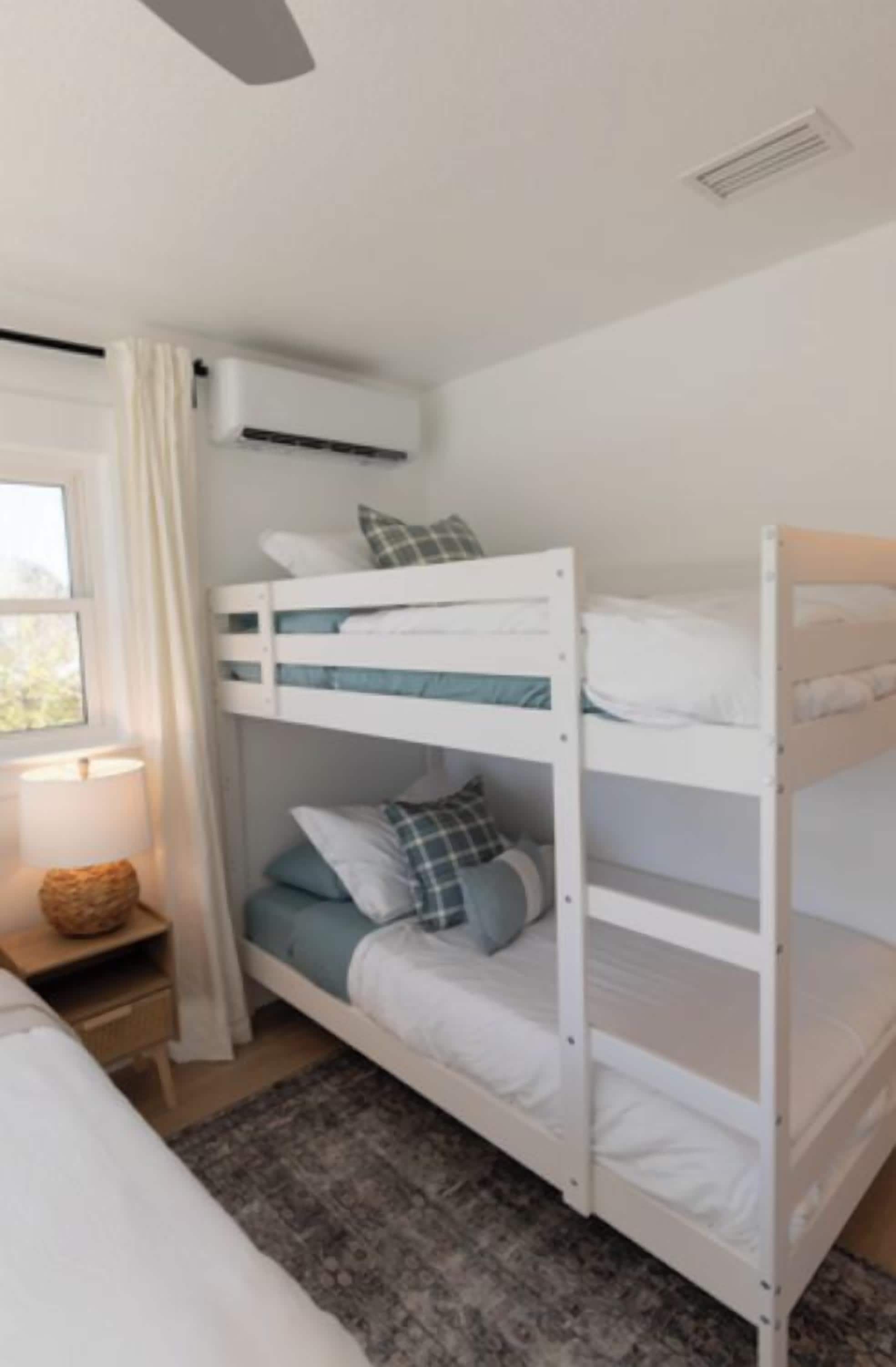 A bunk bed with white bedding, a bedside table with a lamp, and a window with curtains.