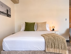 Uma cama com roupa de cama branca, duas almofadas, um cobertor e uma mesa de cabeceira com abajur, além de um quadro na parede.