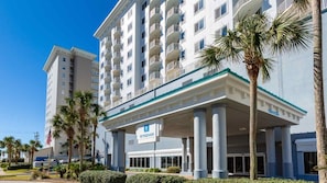 Un hotel de varios pisos con una entrada central, palmeras y un cielo azul despejado.