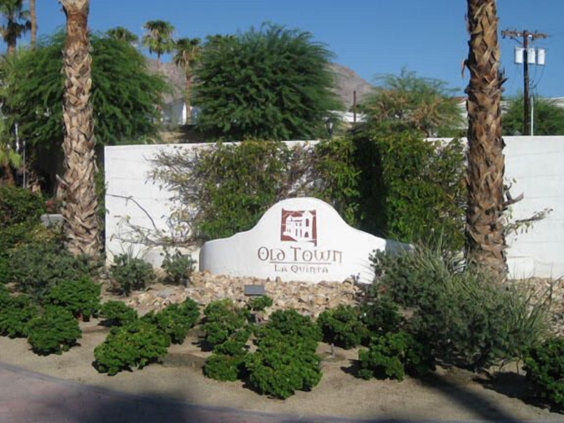 Une enseigne indiquant « Old Town La Quinta », avec un fond blanc et des lettres rouges, entourée de verdure et de palmiers.