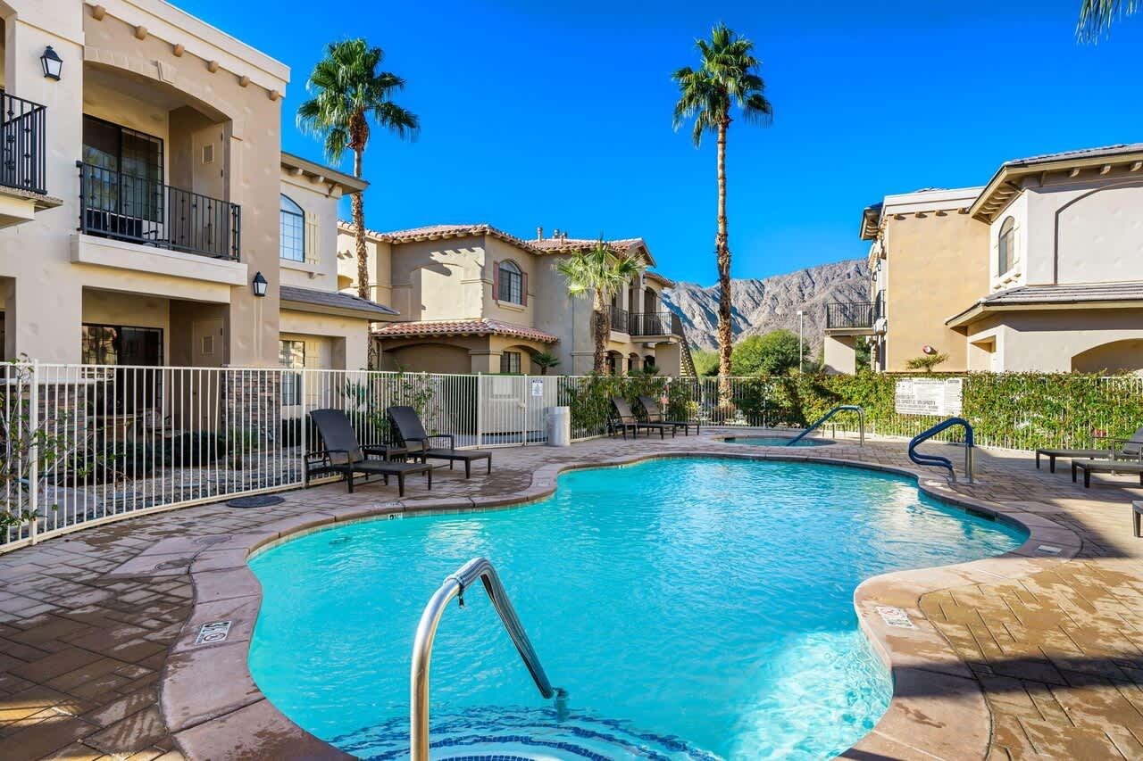 Una piscina de borde curvo, rodeada por una cerca blanca, y junto a un edificio con balcones y palmeras.