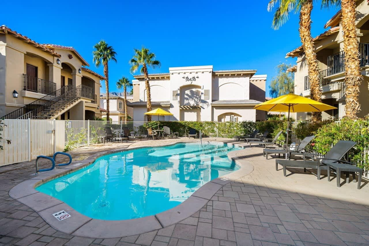 A swimming pool with lounge chairs and umbrellas, surrounded by palm trees and buildings with balconies.