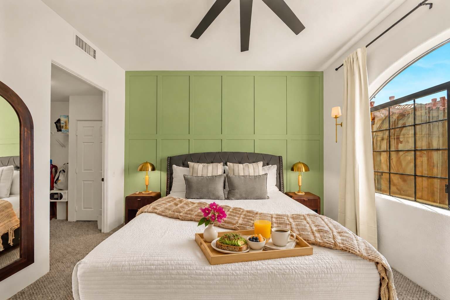 Une chambre d’hôtel avec un grand lit, une tête de lit verte, deux tables de chevet avec des lampes, un plateau avec le petit-déjeuner et une fenêtre donnant sur un beau paysage.