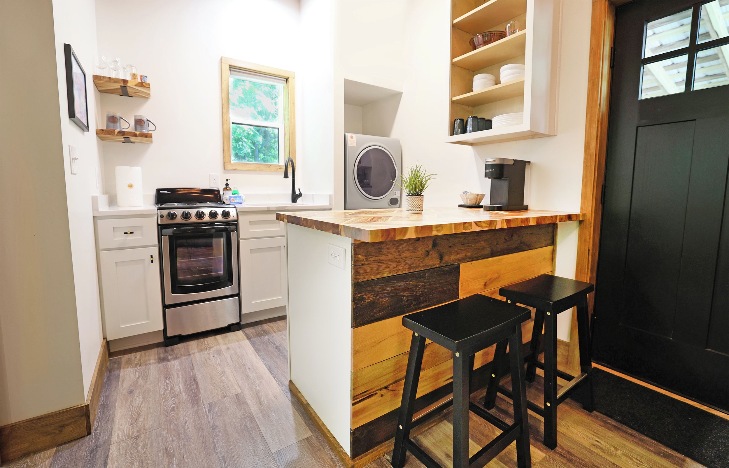 A compact kitchen with a built-in oven, a microwave, and a coffee machine. There are two stools and a small potted plant on the counter.