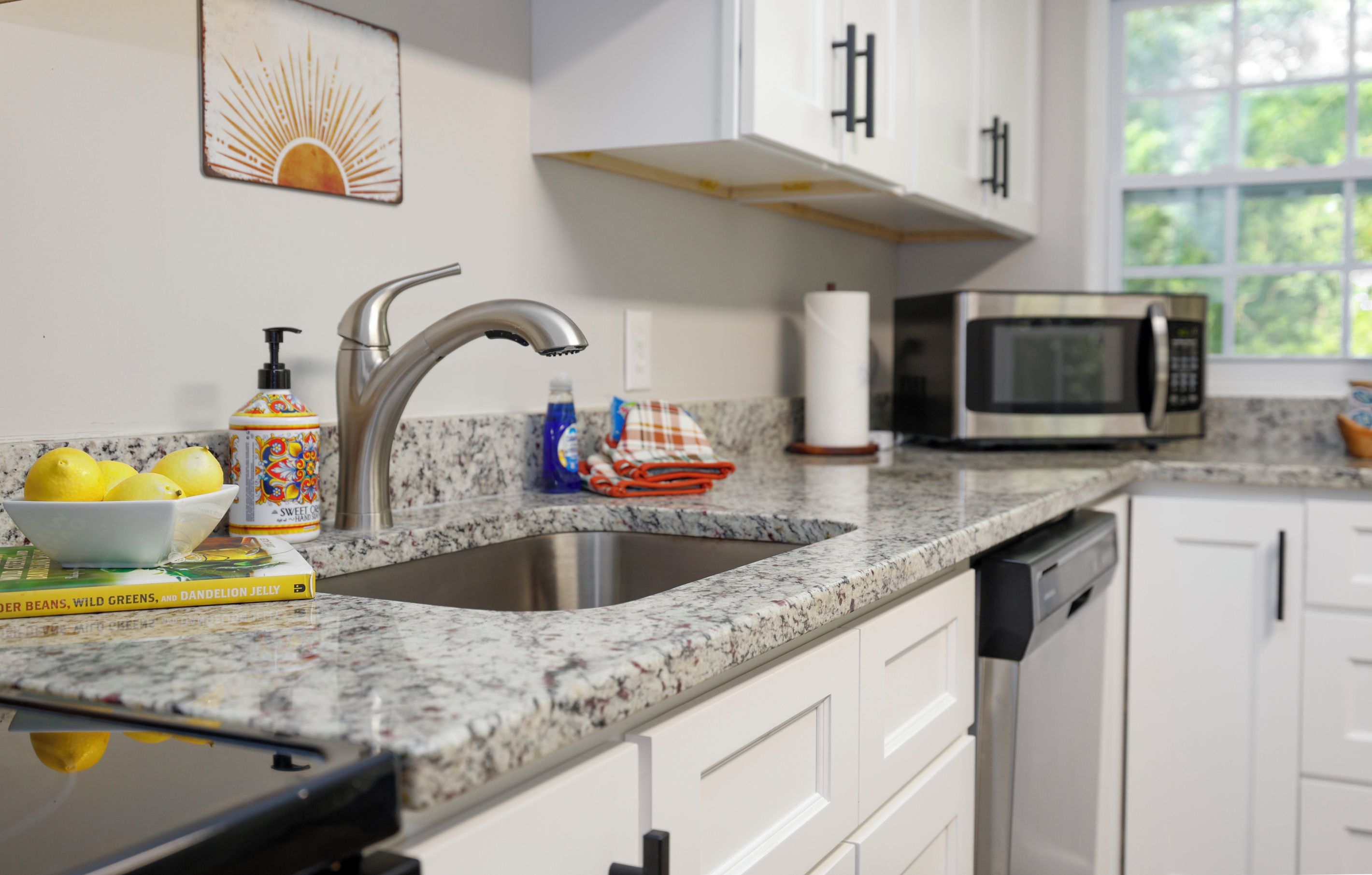 A kitchen with a granite countertop, stainless steel sink, white cabinets, a microwave, and a window with a view of greenery.