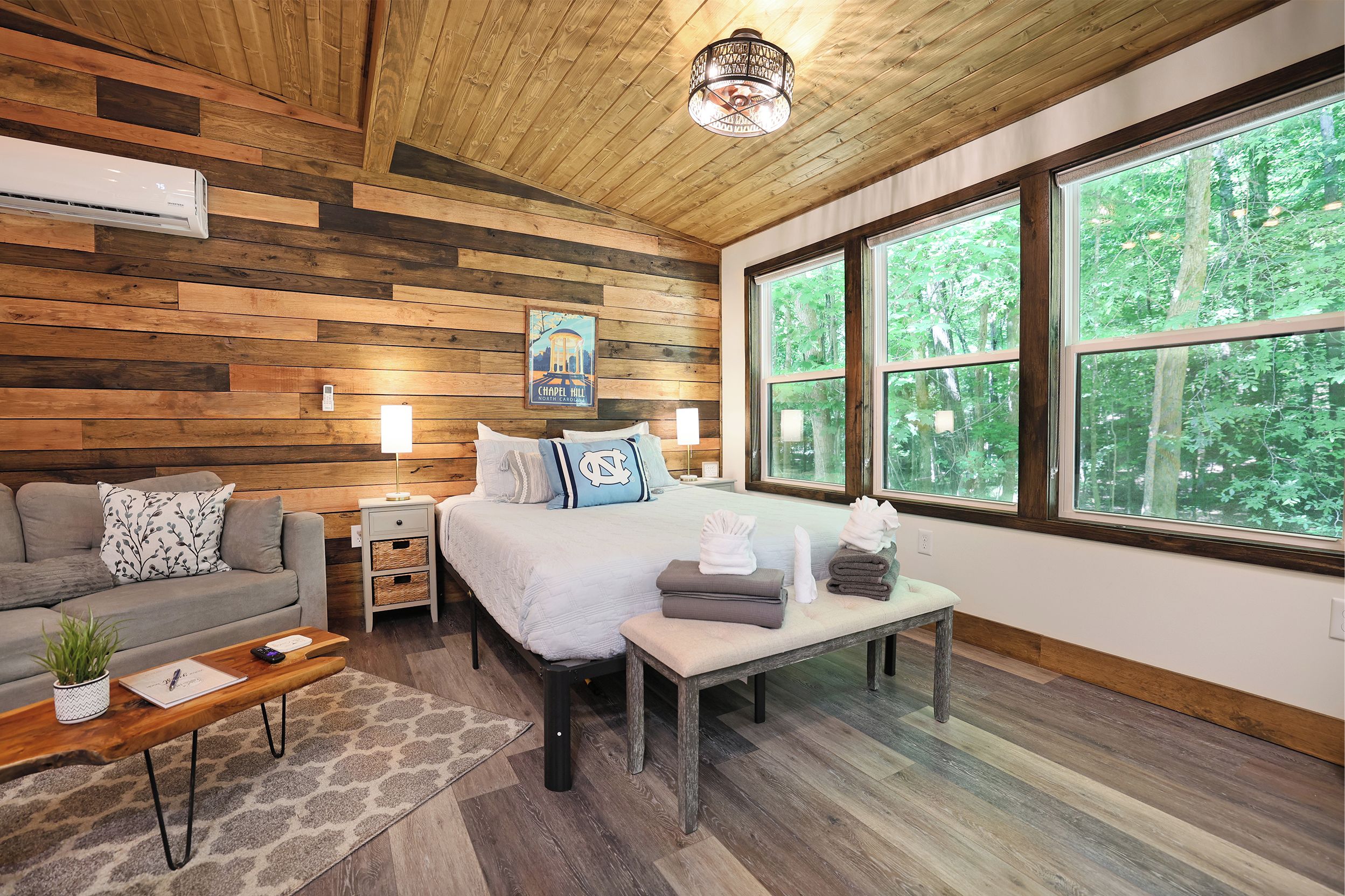 A cozy room with a bed, a sofa, a bench, and a coffee table. There are large windows with a view of trees.