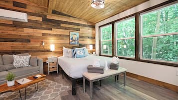 A cozy room with a bed, a sofa, a bench, and a coffee table. There are large windows with a view of trees.