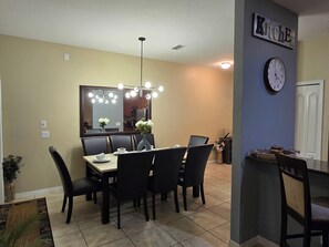 Un coin repas comprenant une table, des chaises, un vase avec des fleurs, une horloge et un panneau indiquant l’emplacement de la cuisine, le tout fixé au mur.