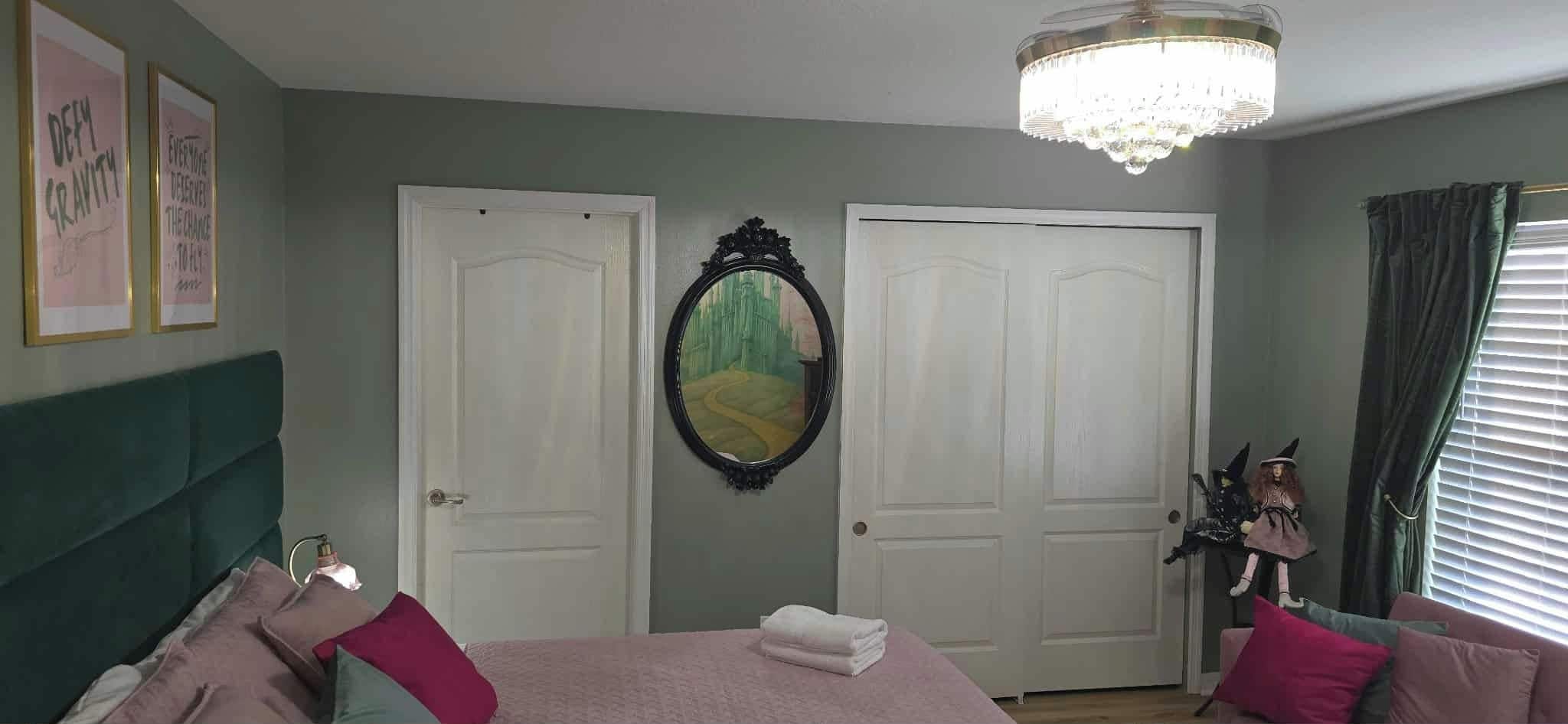 A bedroom with a bed, two doors, a mirror, a chandelier, and a dresser with a doll.
