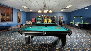 Una mesa de billar con juegos de arcade a su alrededor, una mesa de ping pong y un dardo en una sala con alfombra estampada y paredes azules.