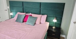 Una cama con cabecera verde azulada, ropa de cama rosa y varios almohadones. Hay dos lámparas de noche en mesitas negras.