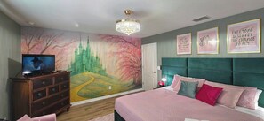 Une chambre à coucher avec un grand lit, une commode, un téléviseur, une fresque murale et des œuvres d’art décoratives aux messages inspirants.
