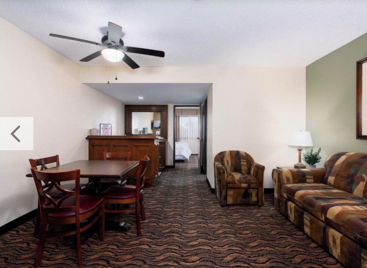 A hotel room with a dining area, a sofa, a chair, a lamp, and a ceiling fan.