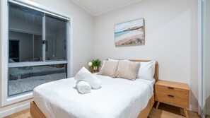 A bedroom with a large window, a bed with white bedding, a bedside table with a lamp, and a painting on the wall. - Hillside Retreat ~ Tranquil 2-bed with mountain views! (Nowra Hill)