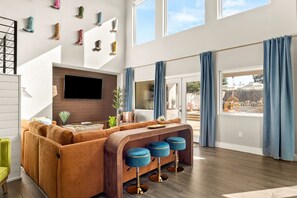 A modern living room with a large TV, a sofa, a coffee table, and a bar area with stools. - Boots & Birdies -Sleeps 22- Luxury Pool+Spa, Pickleball, Golf Simulator, & more (Austin)