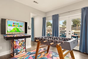 A room with a foosball table, a television displaying a video game, and a colorful rug. - Boots & Birdies -Sleeps 22- Luxury Pool+Spa, Pickleball, Golf Simulator, & more (Austin)