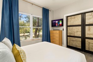 A bedroom with a bed, a TV on the wall, a wardrobe, and a window with curtains. - Boots & Birdies -Sleeps 22- Luxury Pool+Spa, Pickleball, Golf Simulator, & more (Austin)