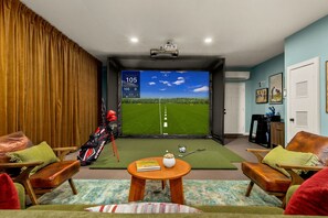 A room with a large screen displaying a golf course, a golf bag, and a table with a book and a decorative item. - Boots & Birdies -Sleeps 22- Luxury Pool+Spa, Pickleball, Golf Simulator, & more (Austin)