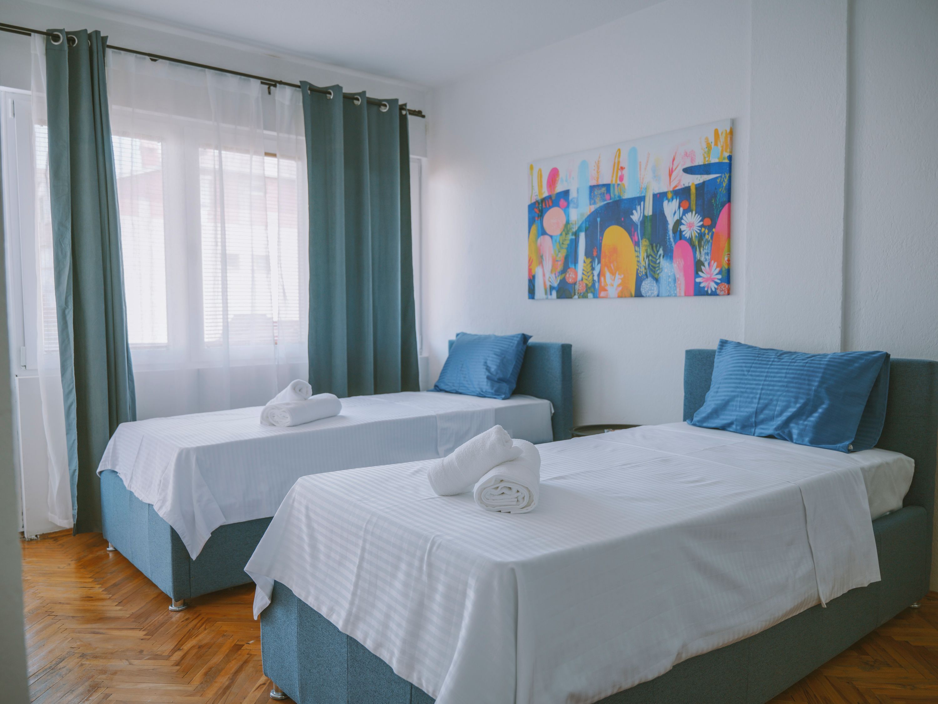 Habitació d'hotel amb dos llits, un gran finestral amb cortines i un quadre abstracte i colorit a la paret.