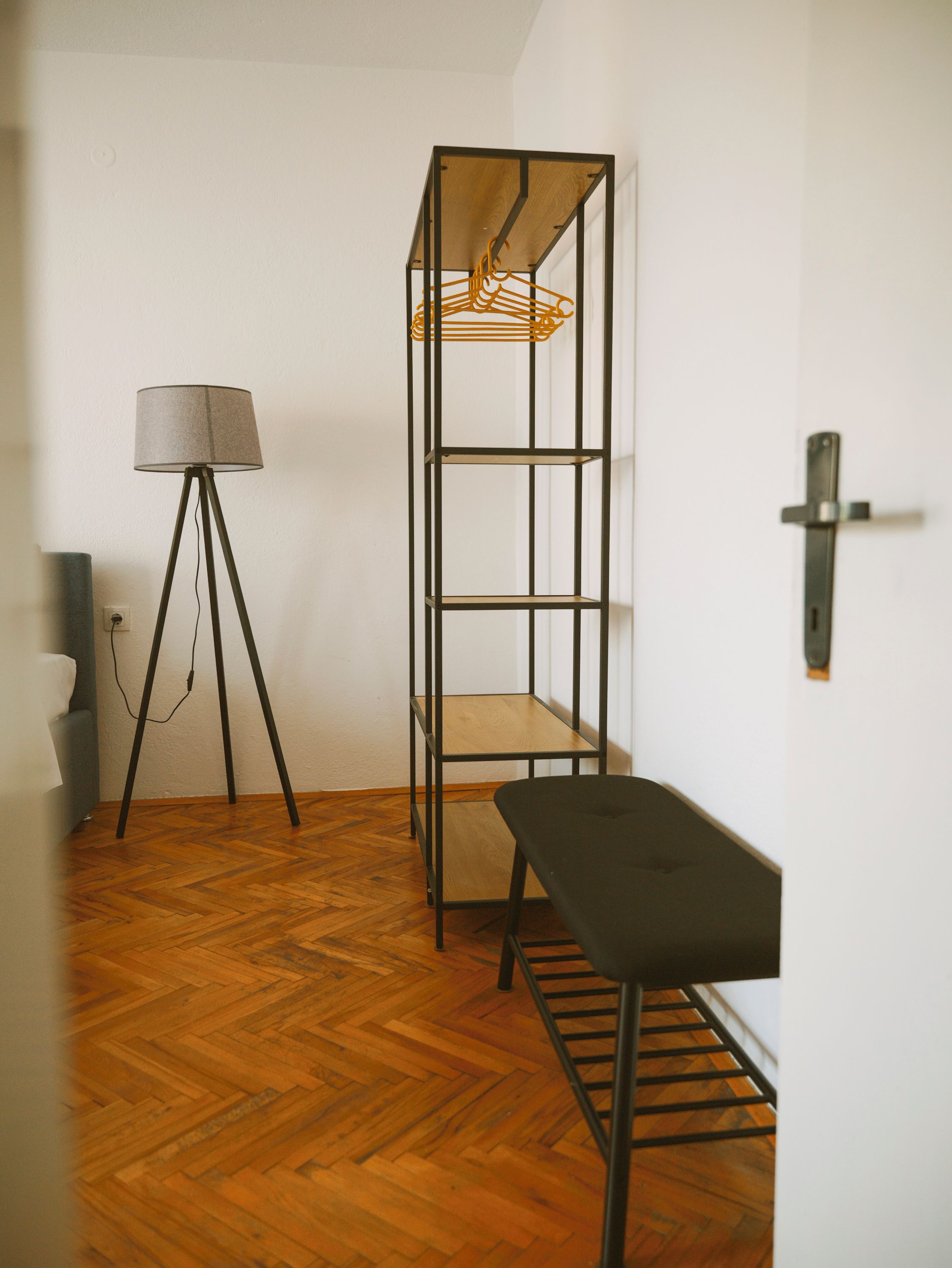 A room with a wooden floor, a metal shelf unit with a hanging rack, a floor lamp, and a bench.
