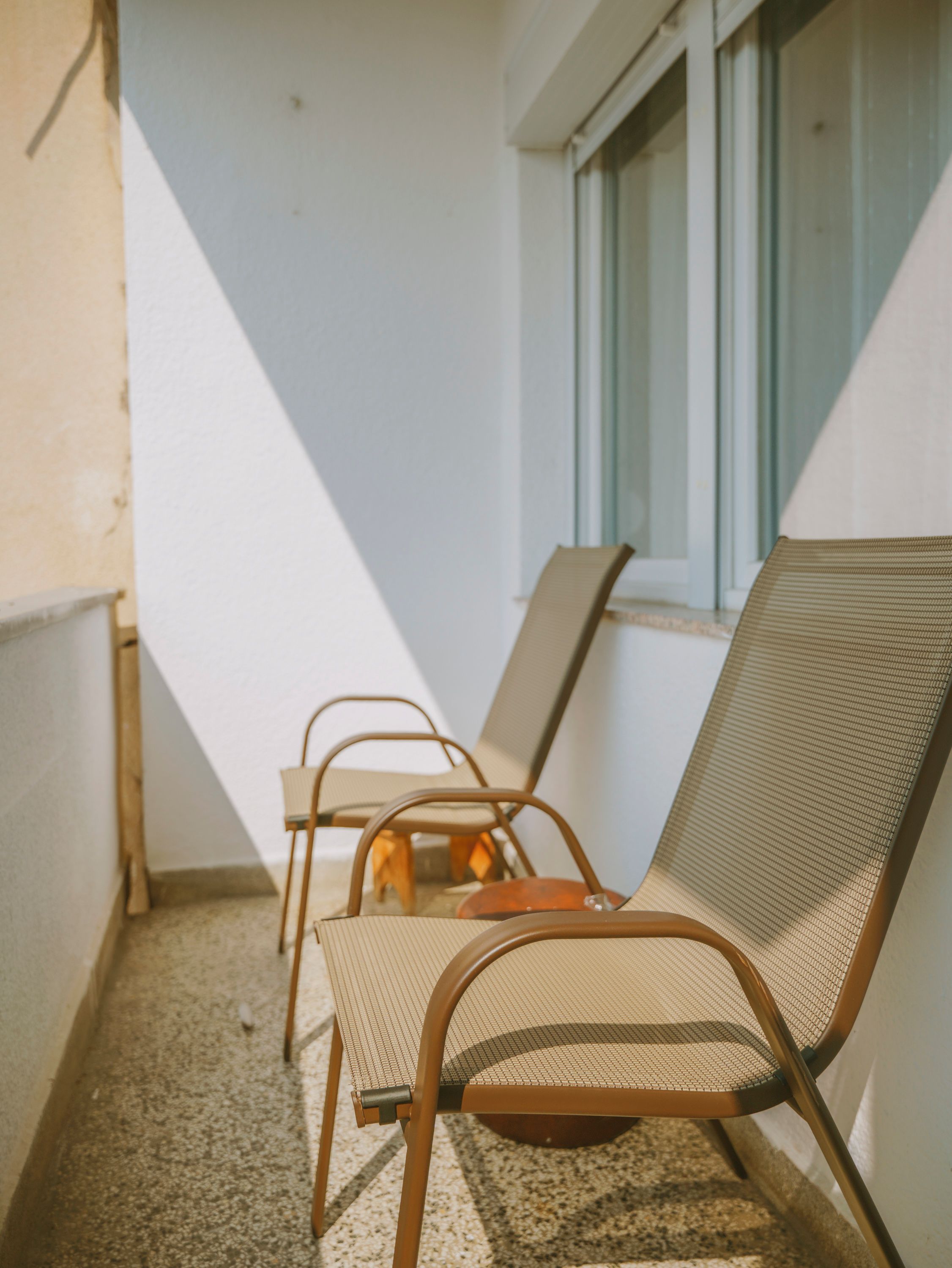 A balcony with two metal chairs and a table, a window, and a white wall.