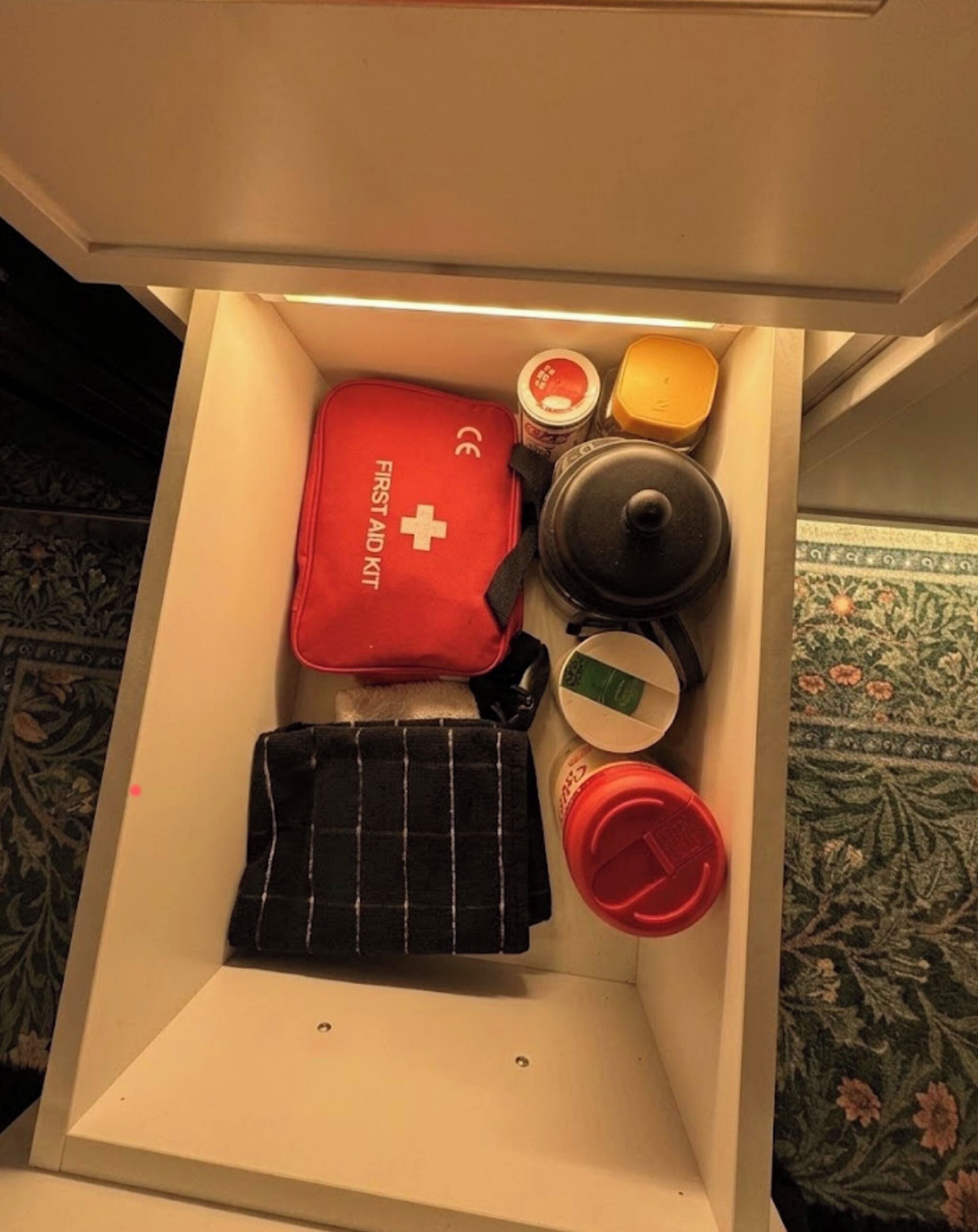 A drawer containing a red first aid kit, a black toiletry bag, a metal container, a glass jar, and a red bottle.