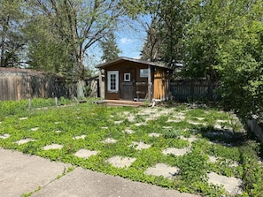 A small wooden cabin with a door and window, surrounded by a garden with various plants and a wooden fence. - Bungalow Rental in Cambridge Comfort and Convenience Await (Cambridge)