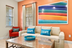 Una sala de estar con un sofá, dos almohadas con la palabra 'ESCAPE', una mesa de centro de vidrio y un cuadro abstracto colorido en la pared.