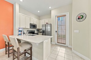 Cocina con armarios blancos, refrigerador de acero inoxidable, microondas y horno empotrado. Hay una ventana con persianas, una puerta con manija y un logotipo en la pared.
