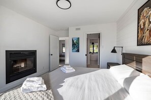 A modern bedroom with a fireplace, a bed, a painting, and a view of a bathroom. - L'Aquarelle : Riverfront Home, 180° View & Hot Tub (Saint-Irénée)