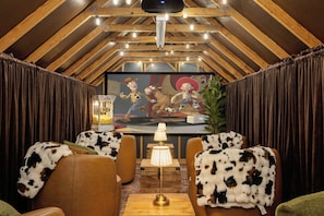 Een gezellige kamer met een hoge plafond, houten balken, een groot schilderij van stripfiguren, een popcornmachine en twee stoelen met koeienprint.