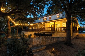 A two-story house with a stone foundation, a wooden deck, and string lights. - Private 10-Acre Estate · Heated Pool · Sleeps 24 (Fredricksburg)
