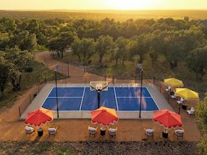 Ein Basketballplatz im Freien mit Sitzgelegenheiten und Sonnenschirmen, umgeben von Bäumen.