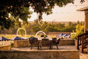 An outdoor patio with a stone table and chairs, surrounded by umbrellas and a stone wall. - 40-ft Heated Pool & Spa, Sport Court, Game Room (Fredricksburg)