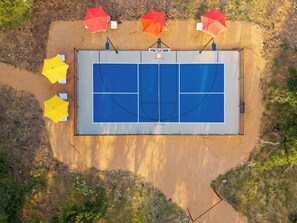 Eine Vogelperspektive auf einen Tennisplatz mit vier Sonnenschirmen, umgeben von sandigem Untergrund und spärlicher Vegetation.