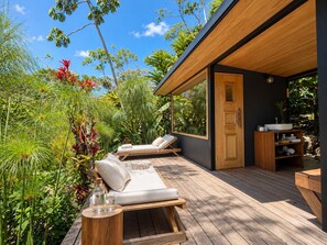 A wooden deck with two lounge chairs and a small table, surrounded by lush tropical plants and a clear blue sky.
