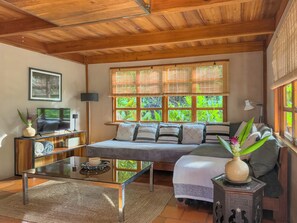 Ein gemütliches Wohnzimmer mit einer Couch, einem Couchtisch und einem Fernseher. Es gibt Fenster mit Jalousien, eine Lampe und eine Vase mit Blumen.
