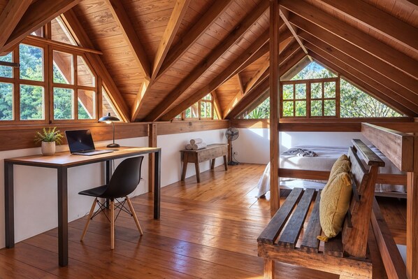 A loft bedroom with a wooden ceiling, a desk with a laptop, a chair, a bed, and a window overlooking greenery.