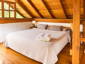 A bedroom with a wooden ceiling, a large bed with a beige bedspread, two pillows, and a nightstand with a lamp. - Casa Nanita | Chirripó riverfront mountain retreat (Perez Zeledon)